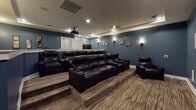 This single-level home theater is thoughtfully designed to deliver an immersive cinematic experience. The room is finished in deep slate-blue walls that set a dramatic, theater-appropriate atmosphere, complemented by crisp white baseboards and a matching raised dividing wall that separates the seating tiers. The flooring throughout features a striped, neutral-toned carpet that adds warmth while reducing sound reflection.

The seating area is arranged in two tiers, with a raised platform at the rear accommodating additional viewers and ensuring unobstructed sightlines to the screen. Dark leather reclining sofas and a matching recliner chair provide comfortable, cinema-style seating with built-in cupholders. A ceiling-mounted projector delivers the picture onto a large, wall-mounted projection screen positioned prominently at the front of the room. Flush in-wall speakers are integrated into the front wall, supporting a surround sound configuration, with additional speaker panels visible along the side walls.

Recessed ceiling lighting and wall-mounted sconces offer layered, adjustable illumination. The entryway features a decorative ledge and cinema-themed wall art, including "Theater" and "Movie Night" signs and film strip artwork, reinforcing the dedicated entertainment purpose of the space.
