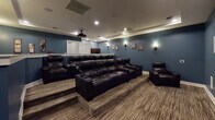 This single-level home theater is thoughtfully designed to deliver an immersive cinematic experience. The room is finished in deep slate-blue walls that set a dramatic, theater-appropriate atmosphere, complemented by crisp white baseboards and a matching raised dividing wall that separates the seating tiers. The flooring throughout features a striped, neutral-toned carpet that adds warmth while reducing sound reflection.

The seating area is arranged in two tiers, with a raised platform at the rear accommodating additional viewers and ensuring unobstructed sightlines to the screen. Dark leather reclining sofas and a matching recliner chair provide comfortable, cinema-style seating with built-in cupholders. A ceiling-mounted projector delivers the picture onto a large, wall-mounted projection screen positioned prominently at the front of the room. Flush in-wall speakers are integrated into the front wall, supporting a surround sound configuration, with additional speaker panels visible along the side walls.

Recessed ceiling lighting and wall-mounted sconces offer layered, adjustable illumination. The entryway features a decorative ledge and cinema-themed wall art, including "Theater" and "Movie Night" signs and film strip artwork, reinforcing the dedicated entertainment purpose of the space.