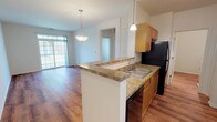 This single-floor residence offers a thoughtfully designed layout that blends comfort and functionality. The open-concept kitchen serves as a central hub, featuring warm honey-toned wood cabinetry, granite-look countertops, a peninsula with a built-in sink, and black appliances including a refrigerator and dishwasher. Pendant lighting above the peninsula adds a decorative touch while illuminating the workspace. The kitchen flows seamlessly into a spacious living and dining area, where hardwood-style flooring extends throughout, creating a cohesive and inviting atmosphere. A sliding glass door in the living area brings in generous natural light and provides outdoor access. The entryway is bright and welcoming, accented by a transom window above the front door. The bedrooms are well-sized, each featuring the same warm flooring, clean white walls, and ample natural light from large windows with outdoor views. A full bathroom includes a soaking tub with tiled surround, a vanity with storage, and consistent flooring that ties the space together. A hallway connects the main living areas to the private rooms, with additional closet and utility access noted throughout. The overall ambiance is clean, neutral, and move-in ready.