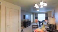 This single-floor residence offers an open-concept layout that seamlessly connects the dining and living areas into one cohesive space. The living room is anchored by a wall-mounted flat-screen television atop a dark-finished media console, while an elegant chandelier with frosted bell-shaped shades serves as a central lighting fixture, adding a decorative touch overhead. A secondary flush-mount ceiling light further illuminates the space. The walls are finished in a cool gray tone, complementing the neutral carpeted flooring throughout. Large windows dressed with two-toned curtains in gray and orange allow generous natural light to fill the room, creating a bright and welcoming atmosphere. The living area accommodates a cushioned seating arrangement centered around a glass-top coffee table. A white-paneled door near the media console provides access to an adjacent area or exterior. The dining space, visible in the foreground, includes dark wood chairs and a table styled with decorative glassware, contributing to the overall warm, casual ambiance of the home.
