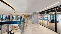 Located at 1000 Vermella Way in New Brunswick, NJ, this expansive amenity floor offers a thoughtfully designed collection of spaces that cater to a wide range of lifestyles. The floor spans a generous footprint and is organized around a striking open corridor with light wood flooring, concrete columns, and curved glass railings that overlook a dramatic double-height atrium below, flooding the space with a sense of openness and architectural elegance.

The state-of-the-art fitness center is a standout feature, divided into distinct zones for cardio and strength training. Large floor-to-ceiling windows invite natural light and offer views of the surrounding neighborhood, while geometric LED lighting arrangements on the exposed ceiling add a modern, energetic atmosphere. Rubber flooring and a full suite of commercial-grade equipment make this a comprehensive workout facility.

The lounge areas exude a sophisticated, hotel-inspired aesthetic, with polished marble-style tile floors, designer pendant lighting, and a curated mix of seating including plush chairs and banquette booths. A fully appointed demo kitchen features sleek cabinetry, a gas range, stone countertops, and a mosaic tile backsplash.

A dedicated children's playroom rounds out the amenities, offering a whimsically decorated space with patterned carpeting, built-in bookshelves, and imaginative wall murals — ideal for younger residents.