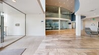 Located at 13900 Tahiti Way in Marina del Rey, California, this single-floor common area space presents a sophisticated, modern design tailored to an upscale coastal setting. The layout encompasses a spacious living/lounge area and a connecting hallway, all finished with large-format stone-look tile flooring that lends a clean, cohesive aesthetic throughout.

The entrance hallway is bright and welcoming, with white walls, recessed lighting, and a hand sanitizer station, leading into the expansive main lounge area. A dramatic curved glass partition wall with a multi-story atrium serves as a centerpiece architectural element, visible from the entry and adding depth and volume to the space. Recessed ceiling spotlights illuminate the curved glass structure, enhancing its visual impact.

The main lounge area is generously proportioned and bathed in natural light from floor-to-ceiling glass doors that open toward a landscaped outdoor area. A defining feature is a wall of illuminated blue glass art panels, evoking the feel of the nearby marina waters and creating a vibrant, immersive focal point. Contemporary seating arrangements — including modular sofas, lounge chairs, and glass coffee tables — are thoughtfully positioned throughout. Curated artwork adorns the walls, reinforcing the polished, gallery-like ambiance of the space. Overall, the design strikes a balance between functionality and refined coastal elegance.