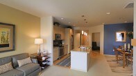This single-floor residence showcases a thoughtfully designed open-concept layout that effortlessly connects the kitchen, dining, and living areas. The kitchen is a standout feature, equipped with stainless steel appliances including a refrigerator, stove, and built-in microwave. Light wood cabinetry extends to the upper walls, offering generous storage, while a white central island with a granite countertop and integrated sink serves as both a functional workspace and a natural gathering point. Pendant lighting above the island adds a warm, inviting glow that complements the recessed ceiling lights throughout. The flooring transitions from warm hardwood in the kitchen to plush carpet in the living area, defining each zone while maintaining a cohesive flow. The living space is cozy and well-lit, with a side table lamp casting a soft ambiance alongside the natural tones of the walls. A hallway leads to additional rooms, hinting at further private spaces beyond the main living area. The overall aesthetic blends contemporary design with comfortable, neutral tones, creating a welcoming and functional environment suited for everyday living.