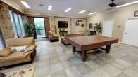 This single-floor community space is designed for relaxation and recreation, offering a well-appointed lounge area that balances comfort with functionality. The room is anchored by a full-size billiards table at its center, making it an inviting hub for social activity. Large tile flooring throughout provides a clean, durable foundation that ties the space together seamlessly.

Seating is generously distributed across the room, with plush armchairs and sofas arranged to encourage conversation and leisure. A wall-mounted flat-screen television is positioned prominently, framed by decorative brick columns that add a warm, rustic architectural character to the otherwise neutral interior. Crown molding along the ceiling adds a refined finishing touch.

Natural light flows in through large windows and a glass door that opens to a lush, green outdoor area, creating an airy and bright atmosphere. Recessed and flush-mount ceiling lighting ensures the space remains well-lit throughout the day and evening. Decorative touches such as potted plants, framed wall art, and accent furnishings contribute to a homey and inviting ambiance. A small desk or writing area along one wall offers a quieter corner for more focused activities. Overall, this space provides a versatile, comfortable environment suited for both casual gatherings and quiet downtime.