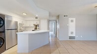 This single-floor residence offers a functional and efficient layout across two bedrooms and two bathrooms. The combined kitchen and living area serves as the heart of the home, featuring light beige tile flooring that extends throughout the space. The kitchen is outfitted with crisp white shaker-style cabinetry, a classic subway tile backsplash, light stone countertops, and a curved peninsula that provides additional prep and seating space. Stainless steel appliances, including a refrigerator, dishwasher, and range, complement the clean aesthetic, and a stacked washer and dryer unit is conveniently integrated into the kitchen area. A hallway connects the main living space to both bedrooms and bathrooms. The bedrooms feature neutral gray walls, tile flooring, ceiling fans with lighting, and windows that invite natural light. The primary bathroom is finished with wood-look tile flooring, a white quartz vanity countertop, and a walk-in shower enclosed by a sleek sliding glass door with tiled walls and a built-in shelf. Throughout the residence, a consistent light and neutral color palette creates a bright, cohesive ambiance, while recessed lighting and ceiling fans contribute to a comfortable and practical living environment.