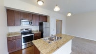 This two-story residence offers a thoughtfully designed interior that blends functionality with modern style. The kitchen serves as a central highlight, showcasing rich dark-wood cabinetry that extends to the upper walls for generous storage capacity. A granite-topped island anchors the space, complete with a double stainless steel sink and pendant lighting overhead, creating an inviting focal point for both cooking and casual gathering. The kitchen is equipped with a full suite of stainless steel appliances, including a refrigerator, electric range with oven, and an over-the-range microwave. Warm hardwood-style flooring runs through the kitchen area, complementing the earthy tones of the cabinetry and countertops. The open layout allows the kitchen to flow naturally into the adjacent living area, which features plush carpeting underfoot. Neutral-toned walls and flush-mount ceiling lighting keep the space bright and versatile. The two-floor layout provides a sense of spaciousness and separation between living and private areas, making this home well-suited for a variety of lifestyles. Clean architectural lines and quality finishes throughout contribute to an overall ambiance that is both practical and polished.