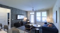 This thoughtfully designed single-floor residence blends contemporary style with everyday comfort. The living area serves as the centerpiece of the home, showcasing a striking marble-patterned accent wall in deep blue and grey tones that anchors the room with dramatic visual impact. A large flat-screen television is mounted against this feature wall, complemented by a sleek dark media console adorned with decorative accessories and warm table lamps that add ambient lighting to the space. The seating arrangement is both stylish and functional, featuring a navy blue sofa, a pair of mid-century modern accent chairs, and a round white coffee table atop a patterned area rug, creating a cohesive and inviting conversation area. A ceiling fan with a brushed metal finish is centrally positioned, providing air circulation while adding an understated modern touch. Natural light floods the room through multiple large windows fitted with horizontal blinds and elegant draping curtains, offering privacy without sacrificing brightness. Light wood-tone flooring runs throughout, providing a warm contrast to the cooler tones of the furnishings. A glass-panel door leads to an outdoor area, further enhancing the sense of openness and connectivity to the exterior.