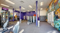 Located at 109 Anderson Hwy in Clemson, SC, this single-floor fitness facility offers a well-appointed and energetic workout environment across a generously sized open floor plan. The space is divided into two distinct zones, each thoughtfully designed to serve different training needs.

The main strength and machine area is defined by bold purple accent walls and structural columns, a drop-panel ceiling with recessed fluorescent lighting, and durable carpet-tile flooring. This zone houses a wide array of selectorized weight machines, free weight benches, a dumbbell rack, and cable systems, all arranged to maximize usable floor space. A large wall mirror enhances the sense of openness, while built-in shelving units provide additional storage. A striking, large-scale tropical mural — featuring tigers, toucans, and lush jungle foliage — serves as a dramatic focal point and adds vibrant character to the space.

The secondary zone features light hardwood-style flooring and a clean white finish, accommodating cycling bikes, medicine balls, and functional training equipment. Wall-mounted televisions and floor-to-ceiling windows along the perimeter flood both areas with abundant natural light while offering outdoor views of the surrounding Clemson landscape. A dedicated cardio row along the window line features treadmills and elliptical machines. Together, the two areas form a cohesive, fully equipped fitness facility with a dynamic and motivating atmosphere.