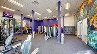 Located at 109 Anderson Hwy in Clemson, SC, this single-floor fitness facility offers a well-appointed and energetic workout environment across a generously sized open floor plan. The space is divided into two distinct zones, each thoughtfully designed to serve different training needs.

The main strength and machine area is defined by bold purple accent walls and structural columns, a drop-panel ceiling with recessed fluorescent lighting, and durable carpet-tile flooring. This zone houses a wide array of selectorized weight machines, free weight benches, a dumbbell rack, and cable systems, all arranged to maximize usable floor space. A large wall mirror enhances the sense of openness, while built-in shelving units provide additional storage. A striking, large-scale tropical mural — featuring tigers, toucans, and lush jungle foliage — serves as a dramatic focal point and adds vibrant character to the space.

The secondary zone features light hardwood-style flooring and a clean white finish, accommodating cycling bikes, medicine balls, and functional training equipment. Wall-mounted televisions and floor-to-ceiling windows along the perimeter flood both areas with abundant natural light while offering outdoor views of the surrounding Clemson landscape. A dedicated cardio row along the window line features treadmills and elliptical machines. Together, the two areas form a cohesive, fully equipped fitness facility with a dynamic and motivating atmosphere.
