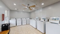 Located at 5055 Harbour Lake Dr in Goose Creek, South Carolina, this single-floor communal laundry facility is designed to serve residents with efficiency and convenience. The space is thoughtfully laid out to accommodate a generous number of top-loading washing machines and dryers, arranged in organized rows along the walls to maximize usability. Light neutral-gray walls and large-format beige tile flooring give the room a clean, bright aesthetic throughout.

Overhead recessed lighting, supplemented by a ceiling fan, ensures the space remains well-lit and comfortable during use. A long, dark stone-topped folding counter runs along one side of the room, providing ample surface area for sorting and folding laundry. A natural wood bench offers a casual seating option for those waiting.

The facility is equipped with coin or card-operated machines and includes a bulletin board for community notices. Natural light filters in through a double-hung window, offering a view of the surrounding greenery and parking area. Safety features include a fire extinguisher and security camera, as well as clearly marked exit signage. The overall ambiance is practical yet well-maintained, reflecting a commitment to providing residents with a clean, functional, and accessible shared laundry experience.