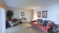 This single-floor residence offers a traditionally styled living room as its central feature. The living room is finished with neutral-toned walls and wall-to-wall carpeting, creating a warm and inviting atmosphere. The space is comfortably proportioned, providing ample room for seating and movement. A coral-toned sofa adorned with plaid and patterned throw pillows serves as the focal point of the room, accompanied by a coordinating plaid armchair that reinforces the classic aesthetic. A decorative console table positioned along one wall holds tasteful ornamental accents, with a framed mirror mounted above it adding depth to the room. Two framed botanical prints displayed side by side on the adjacent wall contribute a refined, traditional touch to the décor. Ambient lighting is provided by a table lamp, enhancing the room's cozy feel during evening hours. The overall ambiance of the space is calm and understated, with a cohesive color palette of muted tones and warm accents that lend the interior a comfortable, livable character well-suited for relaxed everyday living.