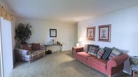 This single-floor residence offers a traditionally styled living room as its central feature. The living room is finished with neutral-toned walls and wall-to-wall carpeting, creating a warm and inviting atmosphere. The space is comfortably proportioned, providing ample room for seating and movement. A coral-toned sofa adorned with plaid and patterned throw pillows serves as the focal point of the room, accompanied by a coordinating plaid armchair that reinforces the classic aesthetic. A decorative console table positioned along one wall holds tasteful ornamental accents, with a framed mirror mounted above it adding depth to the room. Two framed botanical prints displayed side by side on the adjacent wall contribute a refined, traditional touch to the décor. Ambient lighting is provided by a table lamp, enhancing the room's cozy feel during evening hours. The overall ambiance of the space is calm and understated, with a cohesive color palette of muted tones and warm accents that lend the interior a comfortable, livable character well-suited for relaxed everyday living.