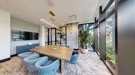 Located at 307 SW 5th St in Fort Lauderdale, FL, this modern conference room offers a sophisticated and thoughtfully designed meeting space. The room's most striking feature is a large, custom-illustrated mural spanning an entire wall, depicting a stylized map of the surrounding Fort Lauderdale neighborhood, complete with palm trees, street names, and local landmarks — adding both character and a strong sense of place to the space.

Floor-to-ceiling glass walls and windows flood the interior with abundant natural light and provide open views of the lush tropical greenery outside, reinforcing the South Florida setting. A substantial natural wood conference table anchors the room, surrounded by plush, curved upholstered chairs in a muted blue-gray tone that complement the space's refined coastal aesthetic.

Exposed concrete columns add an industrial architectural contrast to the otherwise polished interior, while a wall-mounted flat-screen display and a navy blue credenza provide functional amenities for presentations and meetings. Linear LED lighting fixtures overhead deliver clean, even illumination throughout. A tall, statement indoor plant in a white planter near the glass partition introduces a vibrant organic element. Black-framed glass partitions connect this room to adjacent collaborative spaces, maintaining visual openness while defining distinct zones within the broader office environment.