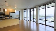 This single-floor space showcases a clean, contemporary design defined by an abundance of natural light. The most striking architectural feature is the wall of floor-to-ceiling windows framed in dark metal, which span the length of the living area and offer expansive outdoor views of the surrounding cityscape and greenery. The open-plan layout seamlessly connects the living area to the kitchen, creating a spacious, versatile environment. Warm hardwood flooring runs throughout, adding a natural contrast to the crisp white walls and ceilings. The kitchen features light wood cabinetry, a stainless steel refrigerator, and a dark-topped breakfast bar that serves as a natural divider between the cooking and living spaces. Pendant lighting above the kitchen adds a refined accent. Structural columns are integrated thoughtfully into the design without disrupting the flow of the space. The large windows also provide access to what appears to be a balcony or outdoor terrace, further extending the living space and enhancing the connection to the outdoors. The overall ambiance is bright, airy, and modern.