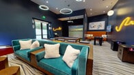 Located at 1825 N Las Palmas Ave in the heart of Hollywood, this single-floor entertainment lounge is a thoughtfully designed recreational space packed with amenities. The room is anchored by deep navy blue walls that create a vibrant, immersive atmosphere, complemented by a wave-patterned carpet and a mix of circular halo pendant lights and recessed ceiling fixtures that set a modern, energetic tone.

At the center of the space sits a full-size pool table, surrounded by high-top bar seating and counter space ideal for social gatherings. A standout feature is the golf simulator, complete with a turf putting surface and a large projection screen, offering an interactive experience for sports enthusiasts. Plush teal velvet sofas with oversized white accent pillows provide comfortable lounge seating throughout.

The space also includes arcade gaming stations, a foosball table, a racing simulator chair, and wall-mounted flat-screen TVs, ensuring a wide variety of entertainment options. Decorative neon light art adds a playful, artistic touch to the walls, while large glass doors along one side allow natural light to filter in. The overall ambiance blends upscale design with a fun, social atmosphere well-suited for recreation and community engagement.