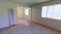 This single-floor space presents a straightforward, functional layout centered around a carpeted living area that connects openly to an adjoining room. The main living space is finished with light beige carpet and neutral sage-green walls, creating a calm, understated atmosphere throughout. Two windows dressed with vertical blinds allow natural light to filter into the room, keeping the interior bright and airy.

A front entry door is centrally positioned along one wall, providing direct access to the living area. To the left, a wall-mounted heating unit offers a practical climate control solution. The open passageway leading into the adjoining room — illuminated by a ceiling-mounted light fixture — suggests a seamless flow between spaces, ideal for flexible use as a dining area or additional living space.

The textured ceiling adds a subtle architectural detail consistent with the overall modest and practical design of the unit. The combination of neutral tones, carpeted flooring, and ample natural light gives the space a clean, livable feel suitable for a variety of furnishing arrangements.