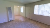 This single-floor space presents a straightforward, functional layout centered around a carpeted living area that connects openly to an adjoining room. The main living space is finished with light beige carpet and neutral sage-green walls, creating a calm, understated atmosphere throughout. Two windows dressed with vertical blinds allow natural light to filter into the room, keeping the interior bright and airy.

A front entry door is centrally positioned along one wall, providing direct access to the living area. To the left, a wall-mounted heating unit offers a practical climate control solution. The open passageway leading into the adjoining room — illuminated by a ceiling-mounted light fixture — suggests a seamless flow between spaces, ideal for flexible use as a dining area or additional living space.

The textured ceiling adds a subtle architectural detail consistent with the overall modest and practical design of the unit. The combination of neutral tones, carpeted flooring, and ample natural light gives the space a clean, livable feel suitable for a variety of furnishing arrangements.