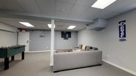 This lounge is a well-appointed gathering space designed for relaxation and entertainment. The room features neutral gray walls that create a calm, cohesive atmosphere, complemented by plush wall-to-wall carpeting in a matching tone. A large gray sectional sofa serves as the centerpiece, positioned to face a wall-mounted flat-screen television, making it an ideal setting for casual viewing or socializing. The drop ceiling is fitted with recessed panel lighting that provides bright, even illumination throughout the space. A single white structural column is centrally located within the room, adding an architectural element without significantly disrupting the open layout. The walls are accented with subtle decorative touches, including a pennant flag, lending a personalized feel to the otherwise understated design. Ductwork is partially visible near the ceiling in one corner, characteristic of a finished lower-level space. The overall aesthetic is modern and functional, with a clean, minimalist approach that keeps the focus on comfort and usability. The space is well-suited for lounging, watching television, or hosting small gatherings.