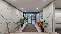 This building lobby and hallway presents a well-designed, contemporary entrance space that makes a strong first impression. The entry vestibule is defined by striking geometric panel detailing on the flanking walls, finished in a soft sage and white palette that lends a clean, modern aesthetic. Floor-to-ceiling glass double doors flood the space with natural light and offer clear views to the outdoor streetscape, creating a seamless visual connection between interior and exterior.

Large-format light-toned tile flooring extends throughout, providing a cohesive and durable surface underfoot. A charcoal entry mat anchors the threshold area, while a small seating arrangement featuring warm cognac-colored chairs and a side table offers a welcoming spot for residents or guests. Potted tropical plants add organic warmth and a touch of greenery to the polished surroundings.

Beyond the main entry, the hallway transitions into a functional corridor with recessed lighting overhead, light grey walls, and continued tile flooring. This area features a resident mailbox station, a wall-mounted intercom system, artwork displays, and clearly marked fire doors. A slatted wood accent element near the elevator or stairwell adds a natural texture to the otherwise crisp interior. The overall ambiance is professional, clean, and thoughtfully curated.