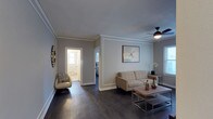 This single-floor residence offers a thoughtfully designed layout with a clean, modern aesthetic. The living area serves as the heart of the space, featuring rich dark hardwood flooring that runs throughout and provides a striking contrast against the soft grey walls. Crown molding adds a refined architectural detail, framing the room with a polished finish. A ceiling fan with a sleek matte black fixture enhances both comfort and style overhead.

The living room is furnished with a contemporary tufted sofa in a warm neutral tone, complemented by a low-profile rectangular coffee table with a metal frame and wood surface. A matching accent chair provides additional seating, while decorative elements such as framed wall art, a sculptural wall accent, and a vase of white orchids contribute to the room's curated ambiance.

Natural light filters in through windows dressed with horizontal blinds, brightening the space with a soft, even glow. An open doorway leads to a hallway connecting to additional rooms, including what appears to be a bedroom and a well-lit bathroom visible in the background. The open sightlines between spaces create a sense of flow and continuity throughout the residence. Overall, the space balances functional design with stylish detailing for a comfortable and inviting atmosphere.