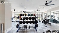 Located at 3007 Petty Rd in Durham, NC, this fitness center offers a comprehensive workout facility spread across a single floor with two dedicated exercise rooms. The space is bright and airy, characterized by large windows that flood the interior with natural light and provide views of the surrounding outdoor greenery. Neutral-toned walls, checkered carpet tile flooring, and multiple ceiling fans contribute to a clean, comfortable workout environment.

The strength training area is generously outfitted with a full power rack system, barbells, weight plates, medicine balls in varying sizes, a tiered dumbbell rack, a heavy bag for boxing, and resistance bands — accommodating a wide range of functional and free-weight exercises. Full-length mirrors line portions of the walls, enhancing the sense of spaciousness and allowing for form monitoring during workouts.

The cardio room features spin bikes, an air bike, treadmills, and a rowing machine, all positioned to take advantage of the natural light from large windows. Recessed lighting and ceiling fans throughout ensure the space remains well-lit and ventilated. The overall layout is thoughtfully designed to support diverse fitness routines, making this facility a well-rounded resource for residents seeking a fully equipped, accessible gym experience.