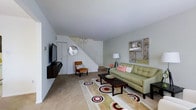 This two-story residence offers a thoughtfully arranged living space that combines functionality with modern style. The main living area serves as the focal point of the ground floor, featuring light-toned walls that create a bright, airy atmosphere. A wall-mounted flat-screen television is positioned opposite a green tufted sofa accented with pink throw pillows, complemented by a wooden slatted coffee table and a patterned area rug with bold circular motifs in neutral, red, and black tones. A single accent chair in warm cognac leather adds an additional seating option. Decorative wall art and a circular mirror contribute personality to the space. A staircase with white railings leads to the upper floor, integrating seamlessly into the living area's layout. The flooring transitions between carpet and hard surface, adding visual texture. A floor lamp provides ambient lighting, enhancing the overall warmth of the room. The space presents a clean, well-organized environment suited for everyday living and entertaining.