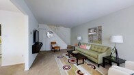 This two-story residence offers a thoughtfully arranged living space that combines functionality with modern style. The main living area serves as the focal point of the ground floor, featuring light-toned walls that create a bright, airy atmosphere. A wall-mounted flat-screen television is positioned opposite a green tufted sofa accented with pink throw pillows, complemented by a wooden slatted coffee table and a patterned area rug with bold circular motifs in neutral, red, and black tones. A single accent chair in warm cognac leather adds an additional seating option. Decorative wall art and a circular mirror contribute personality to the space. A staircase with white railings leads to the upper floor, integrating seamlessly into the living area's layout. The flooring transitions between carpet and hard surface, adding visual texture. A floor lamp provides ambient lighting, enhancing the overall warmth of the room. The space presents a clean, well-organized environment suited for everyday living and entertaining.