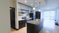 This single-floor space showcases a thoughtfully designed, contemporary interior with a strong emphasis on modern aesthetics. The kitchen serves as the centerpiece, featuring dark espresso cabinetry that extends floor-to-ceiling, complemented by upper glass-front cabinets that add a sense of openness and refinement. Stainless steel appliances, including a freestanding range and a full-size refrigerator, provide a sleek, professional look. A generously sized kitchen island anchors the space, offering an integrated sink and ample countertop space finished in a smooth, neutral-toned surface — ideal for both meal preparation and casual dining. Pendant lighting hangs above the island, adding warmth and a stylish focal point to the kitchen area. Recessed lighting throughout ensures the space is well-lit and inviting. Light hardwood flooring runs underfoot, providing a warm contrast to the darker cabinetry and creating a balanced, harmonious palette. A window allows natural light to filter into the adjoining area, enhancing the bright and airy atmosphere of the open layout. The overall ambiance is clean, modern, and functional — well-suited for those who appreciate contemporary design and practical living spaces.