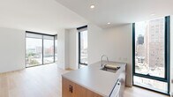 This contemporary 1-bedroom, 2-bathroom residence offers a thoughtfully designed single-floor layout with a clean, modern aesthetic throughout. Light hardwood flooring flows seamlessly across the living spaces, complementing the crisp white walls and creating a bright, airy atmosphere.

The open-concept living and kitchen area is a standout feature, anchored by a stylish kitchen island with a warm wood base and light gray countertop, complete with an integrated sink. Floor-to-ceiling windows with sleek black frames frame sweeping urban views, flooding the space with abundant natural light.

The two living areas offer generous and versatile space, each benefiting from large-format windows that overlook the cityscape. The primary bedroom is a well-proportioned retreat, featuring dramatic corner floor-to-ceiling windows that provide panoramic city views on two sides, with light hardwood flooring continuing throughout.

Both bathrooms are finished with clean, modern fixtures. One features a full soaking tub with white vertical subway tile surround and chrome hardware, while the second includes a tiled walk-in shower, an undermount sink with a light wood vanity, a backlit mirror, and large-format stone tile flooring.

Two hallways provide smooth circulation throughout the residence, and recessed lighting ensures consistent illumination across all areas, contributing to the home's refined and contemporary character.