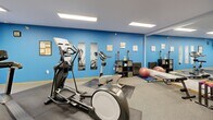 This well-equipped fitness center at 14320 Dysprosium St NW in Ramsey, Minnesota offers a comprehensive workout environment designed to support a variety of exercise routines. The single-floor space is bright and functional, illuminated by recessed fluorescent panel lighting that keeps the entire area well-lit throughout.

The facility features a generous selection of cardio machines, including multiple ellipticals, stationary bikes — both upright and recumbent — a treadmill, and additional cross-training equipment. Protective rubber mat flooring is positioned beneath each machine, providing both safety and stability during use.

One side of the room is accented with bold blue walls and a set of full-length mirrors, creating a visually expansive feel while supporting form-checking during workouts. This area also includes stability balls, a workout bench, and a storage unit stocked with fitness accessories, making it ideal for stretching and supplemental training.

Instructional fitness posters are displayed along the neutral-toned walls, offering guidance for exercise techniques. A wall-mounted HVAC unit ensures year-round climate comfort. The space is accessed through a set of double doors, providing convenient entry and exit. Overall, this fitness center is a practical and well-maintained facility suited for daily cardiovascular and general fitness training.