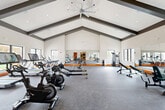 Located at 1 Ridge Rd in Auburn, Massachusetts, this single-floor fitness center offers a generously sized, thoughtfully designed workout facility. The standout architectural feature is the dramatic vaulted ceiling with bold exposed dark beams that rise to a dramatic peak, creating an open, airy atmosphere throughout the main exercise area. A large ceiling fan provides additional airflow, complementing the recessed lighting that keeps the space bright and energizing.

The facility is divided into distinct workout zones. The cardio area lines the perimeter, featuring treadmills, ellipticals, stair climbers, stationary bikes, and rowing machines — many positioned in front of large windows that overlook the outdoor surroundings, allowing natural light to flood in. A dedicated strength training section houses cable machines, weight benches, and a full dumbbell rack with orange-accented stands. Full-length mirrors span entire walls, enhancing the sense of space while allowing users to monitor their form.

The flooring throughout is durable rubber-speckled gym material, with a distinctive orange baseboard accent that adds a vibrant, energetic touch to the otherwise clean, white interior. A separate open studio area with polished floors and wall-mounted TV provides space for stretching or group activity. The entrance corridor leads seamlessly into the main floor, making navigation intuitive and efficient.