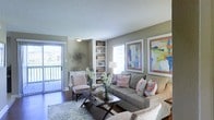 This single-story space centers around a well-appointed living room that exudes a relaxed, coastal-inspired ambiance. The room is finished with warm, neutral-toned walls that provide a versatile backdrop for the colorful, beach-themed artwork displayed prominently above the seating area. A textured ceiling adds subtle architectural interest overhead.

Natural light flows generously into the space through a large sliding glass door that opens directly onto a private balcony, offering outdoor views and enhancing the sense of openness. Vertical blinds provide flexible light control while maintaining the airy feel of the room.

A built-in shelving unit serves as a functional and decorative focal point, offering display and storage space. Dark hardwood-style flooring grounds the room, complemented by a light, patterned area rug that defines the seating arrangement. The living area comfortably accommodates a full sofa, accent chairs, and a glass-topped coffee table. Decorative floral arrangements and coastal accessories complete the inviting, well-curated atmosphere throughout the space.