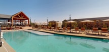 Located at 7800 Crowfoot Valley Rd in Parker, Colorado, this outdoor amenity space is a well-appointed, single-level pool deck designed for relaxation and recreation. The centerpiece is a generously sized, resort-style swimming pool with crystal-clear turquoise water and a smooth curved perimeter, complemented by a separate hot tub for year-round use. Surrounding the pool is a broad concrete deck furnished with rows of cushioned lounge chairs and shade umbrellas, providing ample space for sunbathing and socializing. A covered pergola area anchors one end of the deck and houses a dedicated outdoor kitchen, complete with multiple stainless steel built-in grills set atop tile-clad counters — ideal for community gatherings. Additional seating areas with tables and chairs under umbrellas offer shaded spots for dining and conversation. The space is enclosed by a decorative iron fence with gated access points, ensuring security while maintaining an open, airy feel. The surrounding multi-story residential buildings feature a modern farmhouse aesthetic with board-and-batten siding in neutral and green tones. Panoramic views of the Colorado landscape and wide-open skies enhance the outdoor experience, making this a standout amenity within the community.