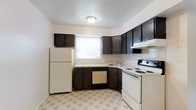 This single-floor space is designed with practicality and functionality in mind. The kitchen serves as a central highlight, featuring dark espresso-toned wood cabinetry that lines the walls in an efficient L-shaped layout, providing ample storage both above and below the countertops. The cabinetry's rich, dark finish contrasts neatly with the light-colored countertops and bright white walls, creating a clean and classic aesthetic. The kitchen is fully equipped with white appliances, including a full-size refrigerator, an electric range with coil burners, a built-in dishwasher, and an overhead range hood, making it a fully functional cooking space. A single window above the sink allows natural light to filter into the kitchen, brightening the workspace. The flooring features a classic geometric patterned vinyl tile in neutral tones, adding a subtle visual interest underfoot while remaining easy to maintain. A flush-mount ceiling light provides consistent overhead illumination throughout the kitchen. The overall ambiance of the space is clean, straightforward, and move-in ready, catering to those who value a well-organized and efficient layout.