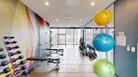 This fitness center offers a thoughtfully designed, single-floor space that accommodates a comprehensive range of workout options. The layout is divided into distinct functional zones, beginning with a cardio area lined with Matrix elliptical machines positioned directly in front of expansive floor-to-ceiling windows. These large-format windows flood the space with natural light while providing sweeping elevated views of the surrounding urban skyline, creating an energizing atmosphere for exercise.

The strength training zone features a full dumbbell rack, kettlebells, resistance bands, TRX suspension straps, foam rollers, and an assortment of stability and medicine balls in vibrant colors. A bold, multicolored geometric mural serves as a striking focal point on one wall, adding visual energy to the space. Large mirrors amplify the sense of openness throughout this section.

A cable machine and additional strength equipment occupy a separate area with more exposed industrial ceiling elements and continued city views. The flooring transitions between light hardwood in the cardio zone and large-format neutral tile in the free weights area. A glass-walled corridor connects to an adjacent outdoor terrace, enhancing the facility's open, airy character. Recessed and pendant lighting complement the abundant natural light throughout.