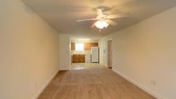 This single-floor space offers a functional and straightforward layout designed for comfortable everyday living. The main living area is finished with wall-to-wall carpeting that adds warmth underfoot, and a ceiling fan with an integrated light fixture provides both air circulation and ambient lighting overhead. Neutral-toned walls throughout lend the space a clean, understated aesthetic.

The living area flows openly into the kitchen, which features warm wood-toned cabinetry providing ample storage space. The kitchen is equipped with a full suite of standard appliances, including a white refrigerator, dishwasher, and a sink positioned beneath a window that allows natural light to brighten the workspace. Light tile flooring in the kitchen area creates a practical, easy-to-clean surface and offers a subtle visual distinction from the carpeted living space.

A doorway visible from the main area leads to additional rooms, suggesting a practical and connected floor plan. Wide baseboards frame the perimeter of the space, adding a simple architectural detail to the overall interior. The open sightlines between the living and kitchen areas create a sense of spaciousness and make the layout well-suited for daily use and easy movement throughout the home.