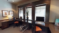 This single-floor workspace is designed with a contemporary aesthetic, combining functionality and style in a compact yet well-organized layout. The room features a built-in dark wood desk that runs along the wall, providing ample surface area for multiple workstations. Two ergonomic office chairs with distinctive orange upholstery add a bold contrast against the darker tones of the furniture and walls.

Three windows fitted with horizontal blinds line the back wall, allowing natural light to filter into the space while maintaining privacy and glare control. The windows are framed by dark wood paneling, lending a polished, professional feel to the room. A decorative potted plant positioned at the center of the desk adds a touch of greenery and visual interest.

Each workstation is equipped with a computer, making the space well-suited for shared or collaborative work. A magazine rack on the floor adds a practical organizational element. The neutral carpet, warm earth-toned walls, and reflective dark desk surfaces work together to create a cohesive, focused environment ideal for productivity.