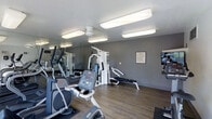 This fitness center offers a practical and well-organized space designed to support a full range of workout routines. The room features wood-look flooring that adds warmth to the otherwise sleek, functional environment, while the gray-toned walls create a clean, modern aesthetic. Large mirrored panels line one wall, visually expanding the space and providing exercisers with proper form feedback during workouts. The facility is equipped with a variety of exercise machines, including an elliptical trainer, a stationary bike, and a multi-station cable and weight machine with an adjustable bench — offering both cardiovascular and strength training options. Overhead, flush-mounted rectangular light fixtures provide bright, even illumination throughout the room, ensuring a well-lit environment at any time of day. A framed informational posting is mounted on the wall, and an additional display screen is affixed near the corner, adding a modern touch. The overall layout is efficient and uncluttered, making it easy to navigate between equipment. This single-floor fitness space is thoughtfully designed to serve users seeking a convenient, all-in-one workout facility.