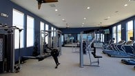This fitness center offers a comprehensive workout environment designed to accommodate a wide range of training styles and fitness goals. The space is defined by striking navy blue walls that create a focused, energizing atmosphere, complemented by light-colored flooring that enhances the sense of openness throughout the room. Recessed lighting is installed across the white ceiling, ensuring consistent, bright illumination throughout, while ceiling fans provide comfortable airflow during intense workouts. Large windows along the perimeter invite abundant natural light, connecting the interior to the outdoors and contributing to an uplifting exercise environment. The facility is outfitted with a robust selection of strength training equipment, including a multi-station cable machine, a Smith machine, a dedicated rowing machine, and various weight racks loaded with plates. Additional resistance and weight machines are arranged throughout the space, offering options for both targeted muscle training and full-body conditioning. Floor-to-ceiling mirrors are strategically positioned to allow users to monitor their form and visually expand the room. The overall layout is thoughtfully organized to allow easy movement between equipment, making this fitness center a fully functional and well-appointed space suited for serious training.