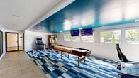 This single-floor game room at 100 Cold Spring Rd in Rocky Hill, Connecticut, delivers a lively, resort-style recreational experience. The space is defined by a striking teal-painted ceiling that adds a bold, energetic character, contrasting with crisp white walls and recessed lighting throughout. A vivid blue, white, and grey geometric-patterned carpet anchors the main gaming area, while neutral large-format tile flooring lines the perimeter hallway, creating a clean visual transition between zones.

The room is equipped with a full-length shuffleboard table as its centerpiece, complemented by a skee-ball lane, a racing arcade cabinet, and a pinball machine, offering a well-rounded entertainment lineup. Two wall-mounted flat-screen TVs are positioned along the windowed wall, providing both ambient viewing and sports coverage. A row of windows along one side brings in natural light and outdoor views, enhancing the open feel of the space.

A standout feature is the expansive floor-to-ceiling glass sliding wall on one side, which offers a direct visual connection to an adjacent indoor pool area complete with lounge seating and pool furnishings. A black-framed glass door provides access to adjoining common areas, making this game room a seamlessly integrated part of a larger amenity complex.