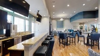This single-floor space is designed as a modern lounge or community gathering area, combining functionality with contemporary style. The open layout seamlessly integrates a bar-height granite counter along one wall, lined with sleek black leather stools featuring nailhead trim detail, making it ideal for casual dining or socializing. Mounted flat-screen televisions are positioned above the counter, enhancing the entertainment-focused atmosphere.

The flooring throughout is warm-toned hardwood, adding a natural contrast to the bold teal and gray color palette that defines the walls. A dedicated dining area occupies the far end of the space, furnished with high-top tables and upholstered chairs in coordinating blue tones. A decorative chandelier hangs above, complemented by a framed wall accent in teal and lime green, adding a focal point to the room.

Recessed lighting runs throughout the ceiling, providing bright, even illumination across the space. Natural light filters in through windows along one wall. The overall ambiance is energetic yet inviting, combining modern design elements with a social, lounge-like atmosphere suited for gatherings and events.