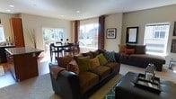 This two-story home offers a welcoming and well-organized layout, highlighted by an open-concept main living area that seamlessly connects the living room and dining space. The living room is generously sized, anchored by a large, dark-upholstered sectional sofa and loveseat adorned with an array of earth-toned accent pillows in olive green, rust, and brown. A plush area rug in neutral tones defines the seating area, while a framed wall mirror and decorative artwork add visual interest to the walls. Recessed lighting in the smooth ceiling provides consistent illumination throughout the space. Large windows dressed with warm terracotta curtains flood the room with natural light and offer views of the surrounding outdoor environment. A sliding glass door leads directly to an outdoor deck, enhancing the indoor-outdoor connection. The dining area sits adjacent to the living room, furnished with a dark wood table and matching chairs, making it functional for everyday meals or entertaining. The overall ambiance is cozy and inviting, characterized by a cohesive warm color palette, practical furnishings, and abundant natural light that make the space both livable and visually appealing.