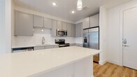 This single-floor residence showcases a clean, contemporary design with a cohesive neutral palette and light hardwood flooring that flows seamlessly throughout. The heart of the home is the open-concept kitchen, which features a generously sized white quartz island that serves as both a prep space and a gathering point. Gray shaker-style cabinetry lines the walls from floor to ceiling, providing ample storage, while a white subway tile backsplash adds a classic touch. Stainless steel appliances — including a French door refrigerator with ice and water dispenser, an electric range, over-the-range microwave, and dishwasher — complete the well-appointed kitchen. Pendant lighting above the island and recessed ceiling lights enhance the bright, airy atmosphere. The bedroom is a calm, light-filled retreat with dual windows fitted with blinds, allowing for natural light and privacy. A ceiling fan adds comfort to the room. The bathroom is equally refined, offering a walk-in tiled shower with a glass door and brushed gold hardware, a gray vanity with a white countertop, and direct access to a walk-in closet with built-in shelving and hanging rods. Throughout the space, white walls and crisp trim maintain a fresh, modern aesthetic.