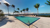This outdoor amenity space centers around a generously sized swimming pool finished with blue mosaic tile detailing, set against a stunning waterfront backdrop. The pool deck is surfaced with light-toned stone pavers that extend comfortably around the perimeter, providing ample space for circulation and relaxation. A row of lounge chairs lines the far edge of the deck, positioned to face the open water, and two large white market umbrellas offer shaded seating options. Tall palm trees are planted throughout the area, swaying in the breeze and reinforcing the tropical character of the setting. The pool is accessible via stainless steel handrails, and the clear, turquoise water reflects the bright blue sky above. Beyond the deck, unobstructed views stretch across a broad waterway, with a distant shoreline visible on the horizon. The overall ambiance is resort-like, combining clean, modern hardscaping with lush natural surroundings. The open-air setting allows for abundant natural light throughout the day, making this a highly functional and visually striking outdoor space suited for leisure and recreation in a warm, coastal environment.