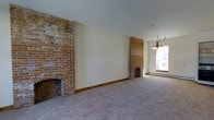 This two-story space blends historic charm with a clean, neutral palette. The main living area is generously proportioned, showcasing two distinct exposed brick fireplaces that serve as the room's defining architectural features. One fireplace anchors the far left wall with a broad, rustic brick surround, while a second, more refined brick fireplace with a arched opening sits closer to the window wall, adding symmetry and warmth to the space. The cream-painted walls contrast beautifully against the rich, red-toned brick, highlighting the craftsmanship of these original elements. Plush neutral carpeting extends across the floor, softening the industrial character of the brick. Natural light filters into the space through a window near the second fireplace, complemented by an elegant multi-arm chandelier that adds a classic finishing touch. Baseboard heating runs along the walls, ensuring year-round comfort. The overall ambiance is one of understated character — a space that honors its architectural heritage while offering a versatile, move-in-ready canvas across two thoughtfully arranged floors.