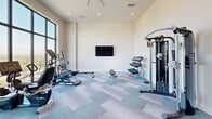 Located at 1250 NW 36th St in Miami, FL, this fitness center offers a well-appointed exercise space designed to support a comprehensive workout experience. The room features soaring ceilings and a striking floor-to-ceiling window wall that floods the interior with abundant natural light while framing expansive views of the Miami skyline and surrounding landscape — an energizing backdrop for any workout.

The space is equipped with a diverse selection of fitness equipment, including cardio machines such as ellipticals, a rowing machine, and a recumbent bike, as well as a multi-function cable machine (Valor AX-5) and a tiered dumbbell rack. A wall-mounted flat-screen television provides entertainment during workouts. The patterned carpet tile flooring in cool blue and grey tones adds a modern aesthetic while providing underfoot comfort.

Recessed lighting and a ceiling fan with a gold accent ensure the environment remains comfortable and well-lit regardless of the time of day. The entry features sleek glass-panel doors with black frames, consistent with the room's contemporary design language. Crisp white walls and clean lines throughout create a focused, distraction-free atmosphere. This fitness center delivers a functional and visually appealing environment for residents seeking an on-site workout facility.