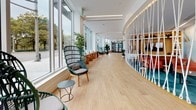Located at 12 Leo M Birmingham Pkwy in Boston's Brighton neighborhood, this contemporary lobby space offers a thoughtfully designed environment that balances open, light-filled areas with distinct zones for gathering and relaxation. The single-floor layout features wide-plank hardwood flooring that runs throughout, contributing to a warm and cohesive aesthetic.

Floor-to-ceiling windows line one side of the space, flooding the interior with natural light and providing clear views of the surrounding streetscape and mature tree canopy. This abundance of natural light enhances the airy, welcoming atmosphere of the lobby.

A striking curved rope partition serves as a central architectural feature, elegantly defining a vibrant lounge area with colorful seating and teal carpeting. On the opposite side of this partition, stylish woven rattan chairs and small side tables are positioned along the window wall, creating intimate seating nooks ideal for relaxing or casual conversation.

Recessed linear lighting in the white ceiling complements the natural light and highlights the clean, modern lines of the architecture. A built-in shelving unit with warm wood backing and LED accent lighting adds a refined, decorative touch, displaying curated books and décor. A bold abstract mural adds an artistic focal point near the corridor, reinforcing the space's creative and contemporary character throughout.