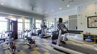 Located at 471 N Hamilton Rd in Gahanna, Ohio, this fitness center offers a well-appointed, single-floor workout facility designed to meet the needs of health-conscious individuals. The space is bright and welcoming, with a row of large windows along the walls that flood the room with natural light, creating an energizing atmosphere for exercise. The ceiling features recessed lighting and a ceiling fan, ensuring the space remains well-lit and comfortable throughout the day. The flooring consists of a patterned, multi-tone carpet tile layout that adds a contemporary aesthetic while providing a cushioned surface underfoot. A lineup of modern cardio equipment, including treadmills and stationary cycling bikes, is arranged in an orderly fashion, all equipped with individual display screens for an enhanced workout experience. Large wall-mounted mirrors on one side of the room amplify the sense of space and allow users to monitor their form during exercise. The overall ambiance is clean, modern, and functional, with soft blue-gray walls contributing to a calm yet motivating environment. This fitness center is thoughtfully designed to offer residents or visitors a convenient and fully equipped space for their daily exercise routines.
