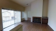 This single-floor residence offers an open and airy living space defined by its impressively vaulted ceilings, which create a sense of grand volume and openness. The main living area is anchored by a classic tile-surround fireplace with a metal screen and glass doors, complemented by a built-in cabinetry unit with a stone-like countertop surface — a functional and visually grounding feature within the room. Warm hardwood-style flooring extends throughout, adding a cohesive and inviting feel underfoot. Large sliding glass doors dressed with vertical blinds line one wall, allowing an abundance of natural light to pour into the space while offering direct access to the exterior. The neutral, light-toned walls provide a clean canvas throughout, enhancing the bright and open ambiance. A textured ceiling adds subtle visual detail overhead. An adjacent room is visible through an open passage, suggesting a well-connected interior layout that allows for easy flow between living areas. The overall atmosphere is open, functional, and ready to accommodate a variety of living arrangements.