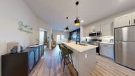 This thoughtfully laid-out one-bedroom, one-bathroom unit offers a cohesive open-concept floor plan that maximizes every square foot. The kitchen is a standout feature, anchored by a central island with a white quartz countertop, stainless steel appliances — including a dishwasher, microwave, and range — light gray cabinetry with matte black hardware, a subway tile backsplash, and stylish pendant lighting overhead. The kitchen flows seamlessly into the living room, which is distinguished by a bold accent wall and light wood-style flooring throughout. French doors lead to a private balcony, filling the space with natural light. The bedroom offers plush carpeting, a ceiling fan, and a large window with views to the exterior. It connects directly to the full bathroom, which features a glass-enclosed walk-in shower with white subway tile, a floating vanity with black hardware, and wood-look flooring. A dedicated walk-in closet with built-in shelving and hanging rods is accessible from the bathroom. An additional closet space accommodates a full-size stacked washer and dryer alongside open shelving for storage. The unit presents a clean, modern aesthetic throughout with consistent finishes and efficient use of space.